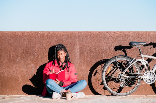 Happy african-american woman with bike sitting on street and writing in a notebook