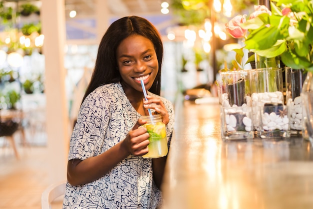 Happy African-American woman with glass of natural lemonade in cafe. Detox drink