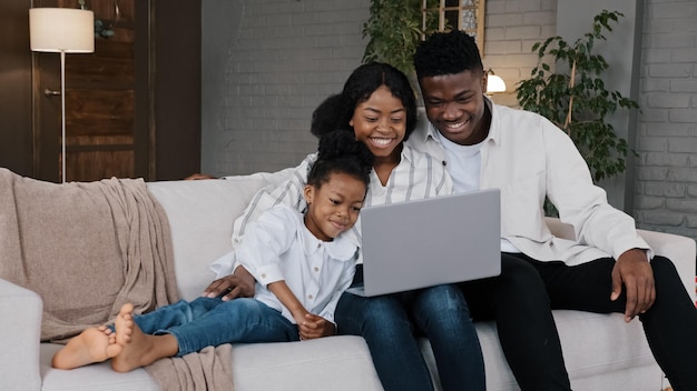 Happy African family and cute kid child girl daughter using laptop looking at computer screen enjoy