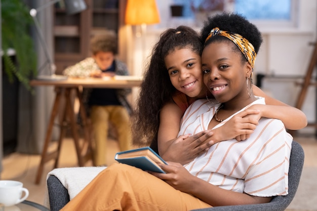 Happy African girl with toothy smile embracing her mother sitting in armchair in front of camera on background of cute boy doing homework