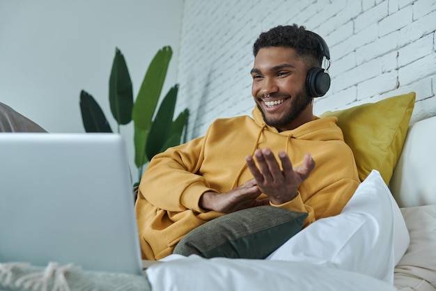 Happy african man in headphones looking at laptop and while lying in bed at home