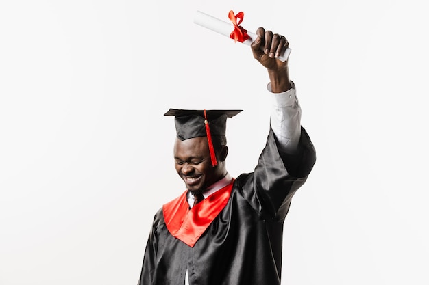 Happy african student in black graduation gown and cap raises masters degree diploma above head on white background Graduate african man is graduating college and celebrating academic achievement