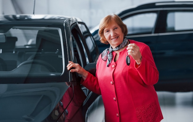 Happy aged woman in formal wear stands in front of modern white car.