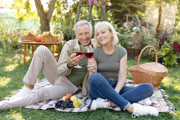 Happy anniversary loving married senior couple drinking wine while sitting on blanket having picnic