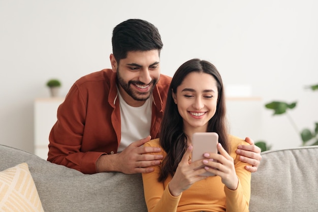 Happy arabic spouses using phone together texting sitting at home