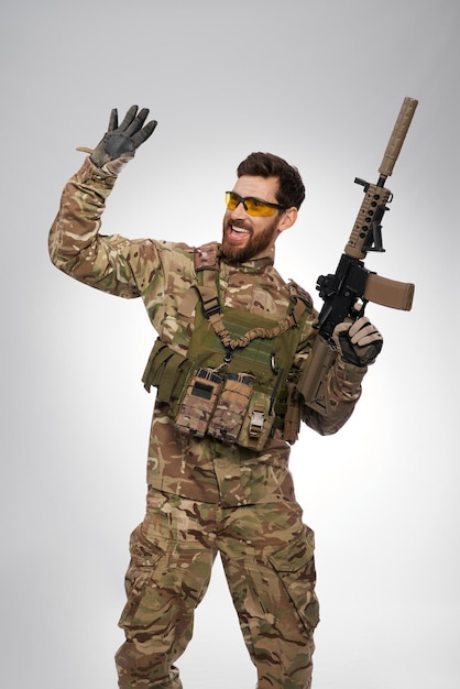 Happy armed man in camouflage outfit waving hand to someone aside