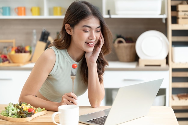 Happy Asia woman eating salad with see computer notebook in kitchen room