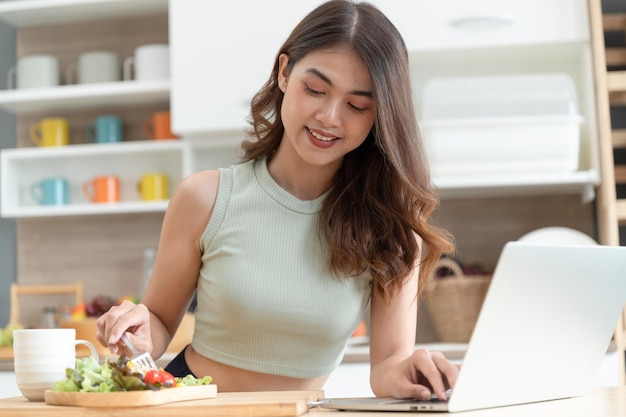 Happy Asia woman eating salad with see computer notebook in kitchen room