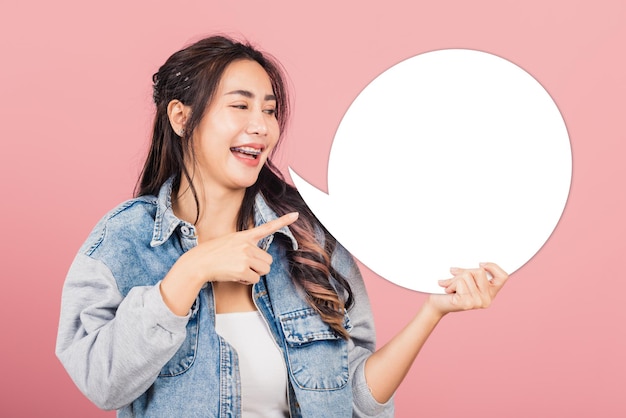 Happy Asian beautiful young woman smiling excited wear denims hold empty speech bubble sign, Portrait female posing finger pointing bubble for your text idea, studio shot isolated on pink background