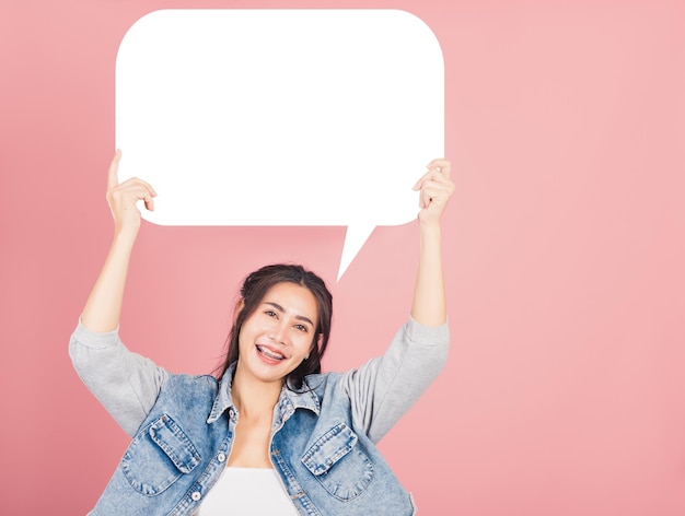 Happy Asian beautiful young woman smiling excited wear denims holding empty speech bubble sign, Portrait female posing show up for your idea looking at camera, studio shot isolated on pink background