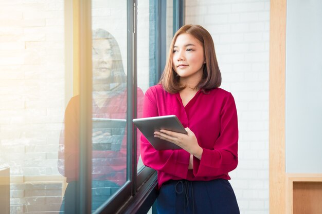 Happy asian business woman standing by large window holding her tablet