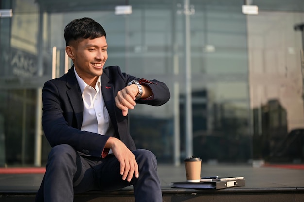 Happy Asian businessman checks the time looking at his watch while sitting on the stairs