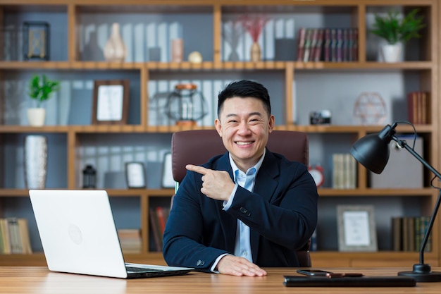 Happy asian businessman showing finger aside sitting in office smiling and looking at camera