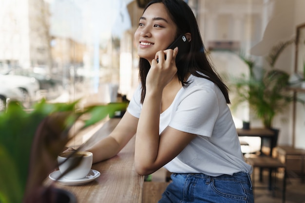 Happy asian candid woman sitting in cafe near the window, looking at street while talking on phone.