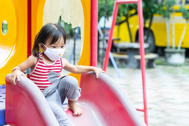 Happy Asian child girl smiling and wearing fabric mask. She playing with slider bar toy at the playground.