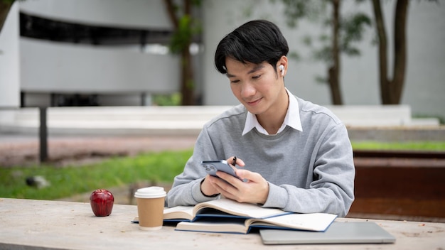 A happy Asian college student is using his smartphone while doing homework at a table in a park