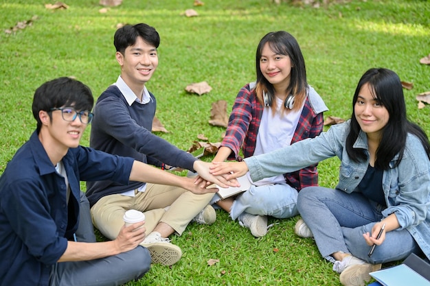 Happy asian college students sitting in the campus's park putting their hands together as a teamwork