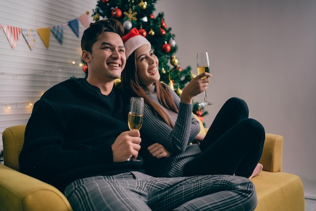 Happy Asian couple cheers and drinks champagne together sitting on the sofa in the Christmas evening