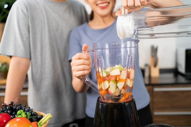 Happy Asian couple enjoys rinsing fresh water in blender machine for making healthy vegan smoothie on the kitchen counter Couple making vegan smoothie together at home for a healthy lifestyle