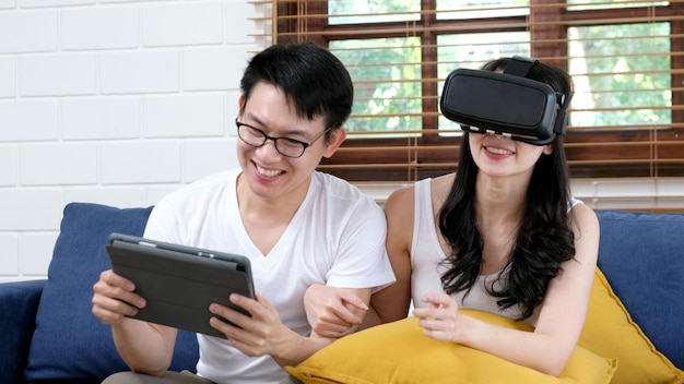 Happy asian couple play virtual reality simulation game while sitting on sofa at home living room, People and technology