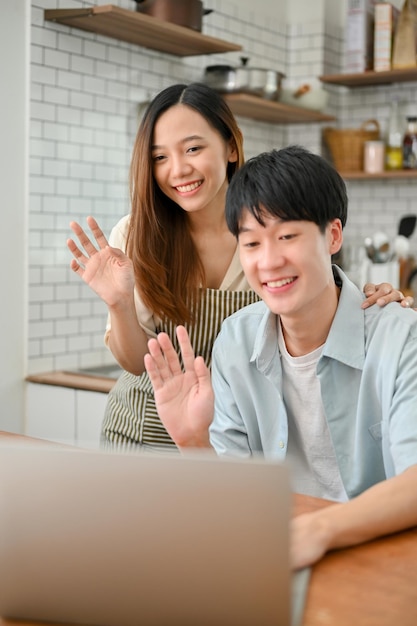 Happy Asian couple saying hi to their parents through a video call on laptop in the kitchen