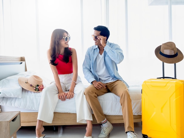 Happy Asian couple in summer young man and woman wearing sunglasses with smile sitting on the bed in the bright bedroom near yellow luggage and beach hat holiday travel summer vacation concept