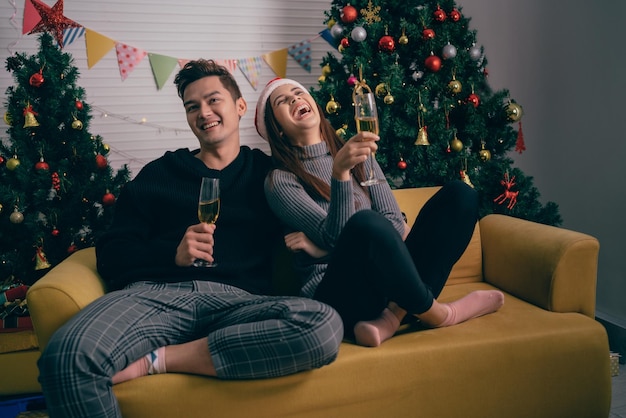 Happy Asian couple toasting champagne together sitting on the sofa in the evening with a Christmas tree and lights in the background Boyfriend and girlfriend at Christmas eve night