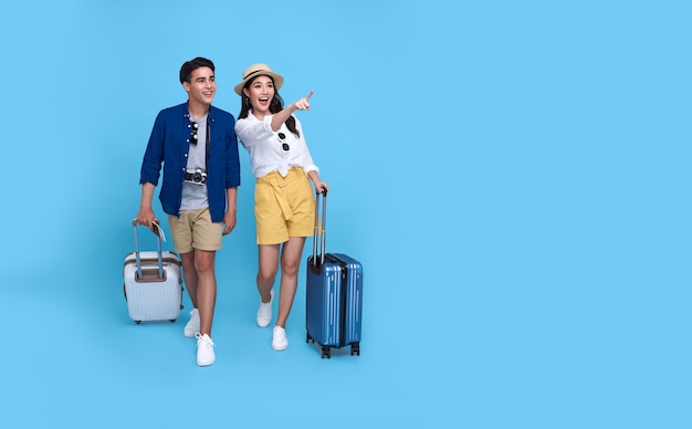 Happy Asian couple tourist hand pointing to copy space with baggage going to travel on holidays isolated on blue.