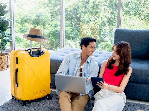 Happy Asian couple young man and woman looking each other while using laptop computer and tablet together for flight booking and trip information in living room near luggage summer vacation