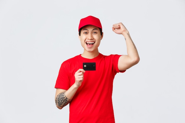 Happy asian delivery man in red cap and t-shirt uniform of carrier company, raising fist and chanting yes, showing credit card. Promo of application, online orders and client purchasing home delivery.