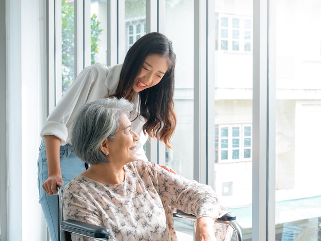 Happy Asian elderly woman mother or grandparents on wheelchair taking care by caregiver smiling young female daughter or grandchild supporting at home on green nature background senior healthcare