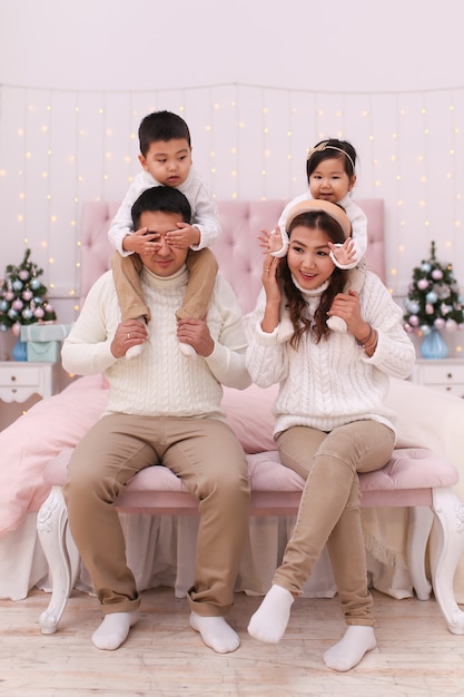 Happy asian family with two children in cozy comfortable sweaters having fun and relaxing on the bed