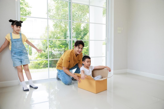 Happy asian father playing with cute small kids daughters and son laughing on moving day boy having fun riding in box in new house