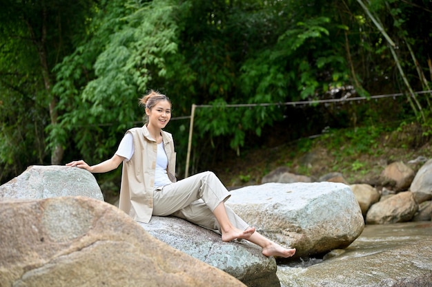 Happy Asian female camper traveler sitting on a river rock near the the river in the forest