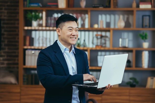 Happy asian freelance businessman working standing holding laptop smiling and rejoicing at home office