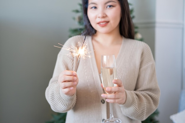 happy asian girl in cozy clothing holding glass of champagne and sparkler on her hands at home