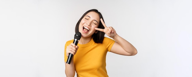 Happy asian girl dancing and singing karaoke holding microphone in hand having fun standing over white background