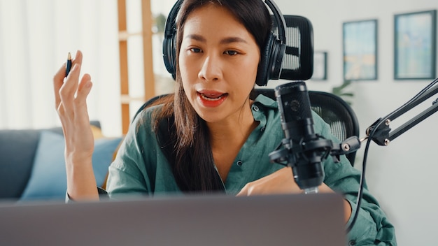 Happy asian girl recording a podcast on her laptop computer with headphones and microphone