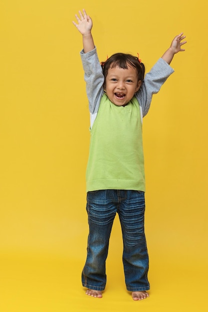 Happy Asian little girl standing and raising hands up isolated over yellow background