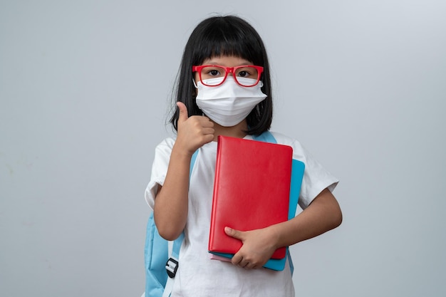 Happy Asian little preschool girl wearing red glasses holding books and red backpack on white isolated background Concept of school kid and education in elementary and preschool home school