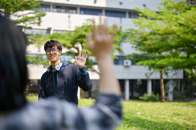 Happy Asian male college student waves his hand to greet his friend at the campus park