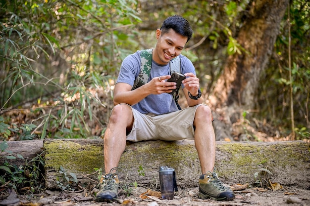 Happy Asian male traveler or trekker using his smartphone while resting on a wooden log