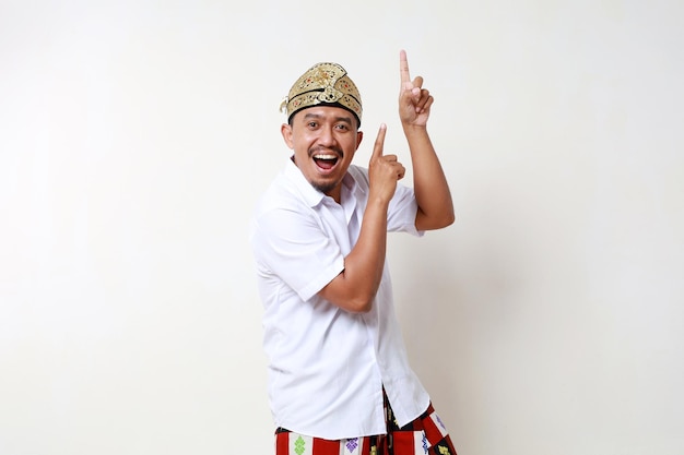 Photo happy asian man in balinese traditional costume standing while looking and pointing above isolated on white background