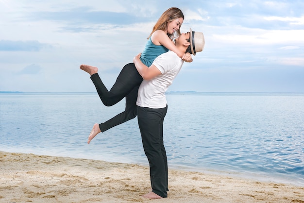 Happy asian man in hat hugging his girlfriend on the beach