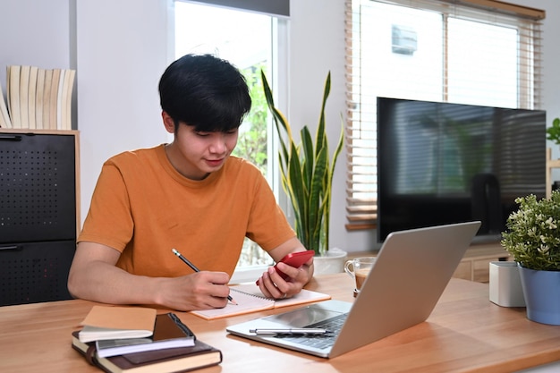 Happy asian man holding smart phone and making notes on notebook.