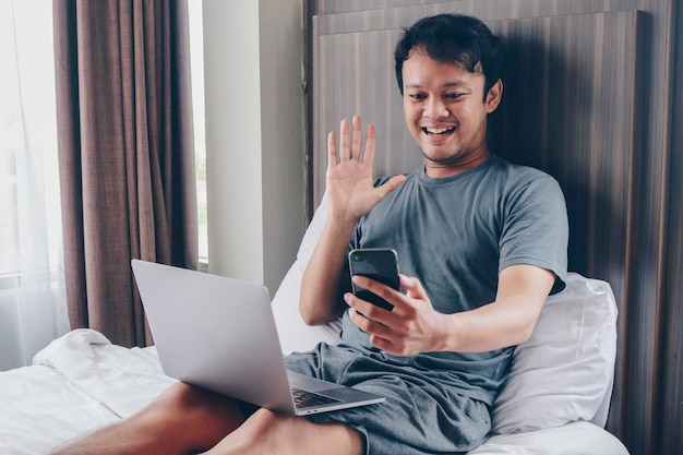 Happy Asian man is working with his laptop with video call in a bed Concept of freelancer successful lifestyle