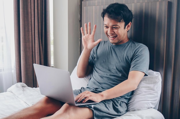 Happy Asian man is working with his laptop with video call in a bed Concept of freelancer successful lifestyle