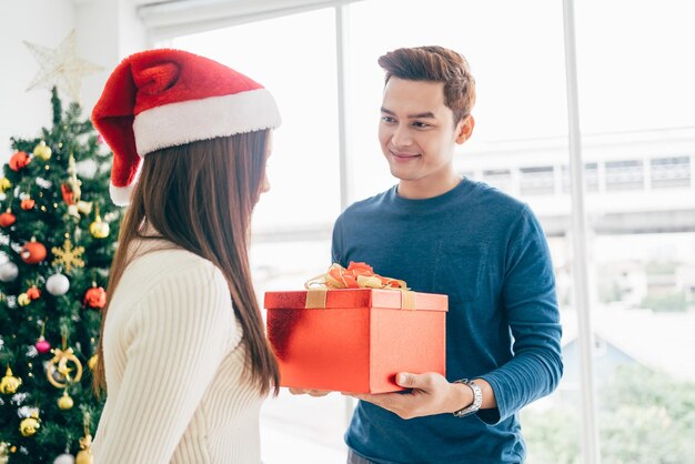 A happy Asian man surprises his girlfriend with Christmas presents at home with a Christmas tree in the background image with copy space