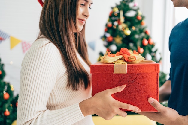 A happy Asian man surprises his girlfriend with Christmas presents at home with a Christmas tree in the background image with copy space