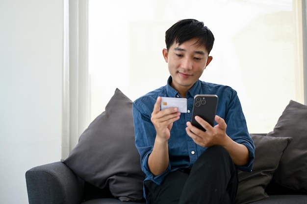 Happy Asian man using a mobile banking to pay his bills while relaxing in a living room